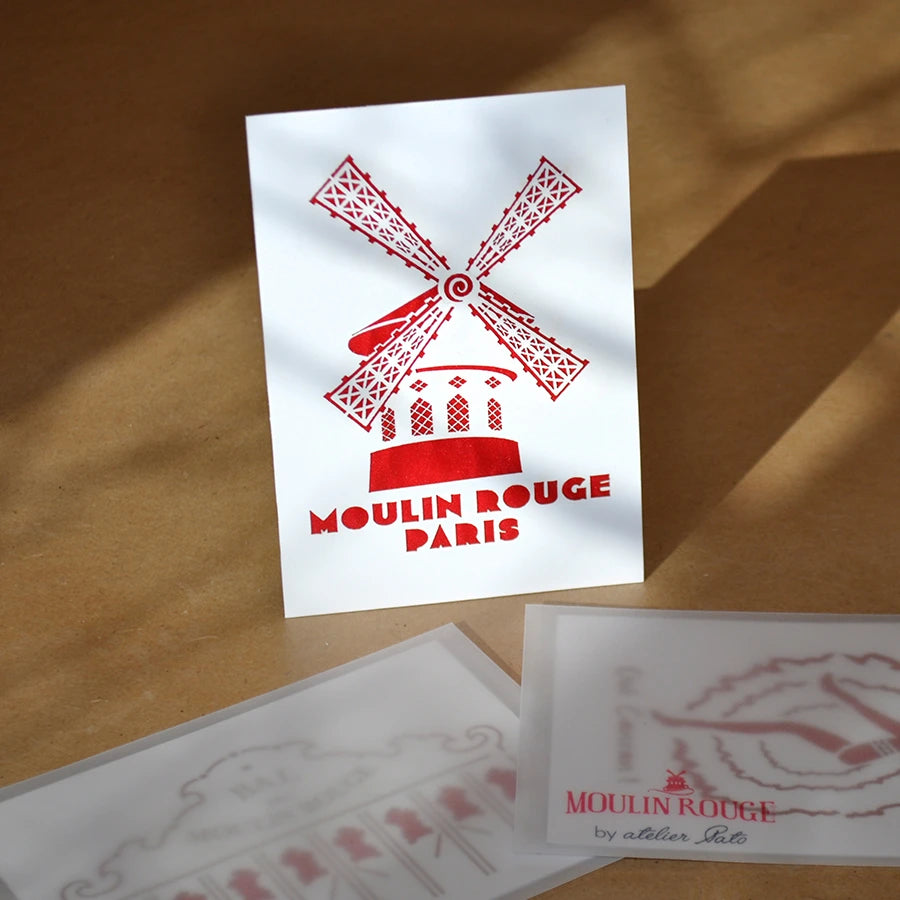 Carte postale blanche et rouge de la façade du Moulin Rouge posée sur une table avec d'autres cartes